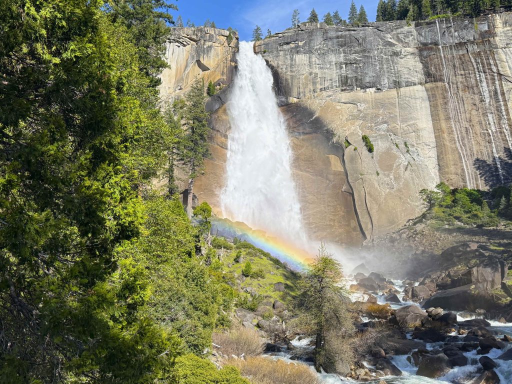 Rainbow forming in the mist at the base of Nevada Fall in Yosemite National Park with rushing water and granite cliffs.