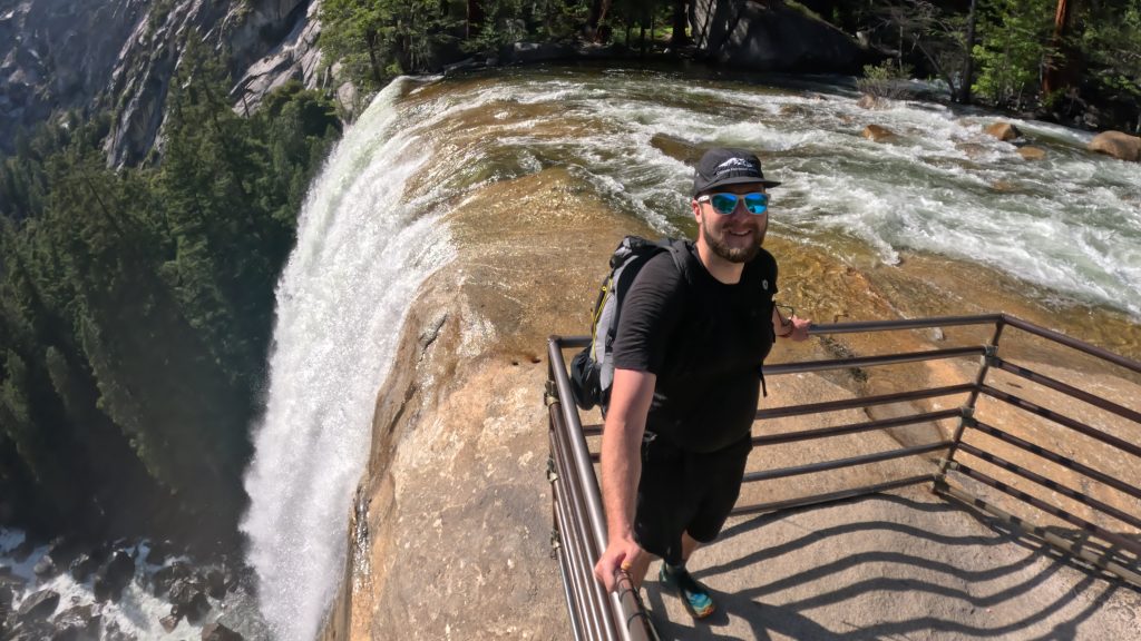 Hiker standing at the railing overlooking the top of Vernal Falls in Yosemite National Park on the Mist Trail with rushing water flowing over the granite cliff.