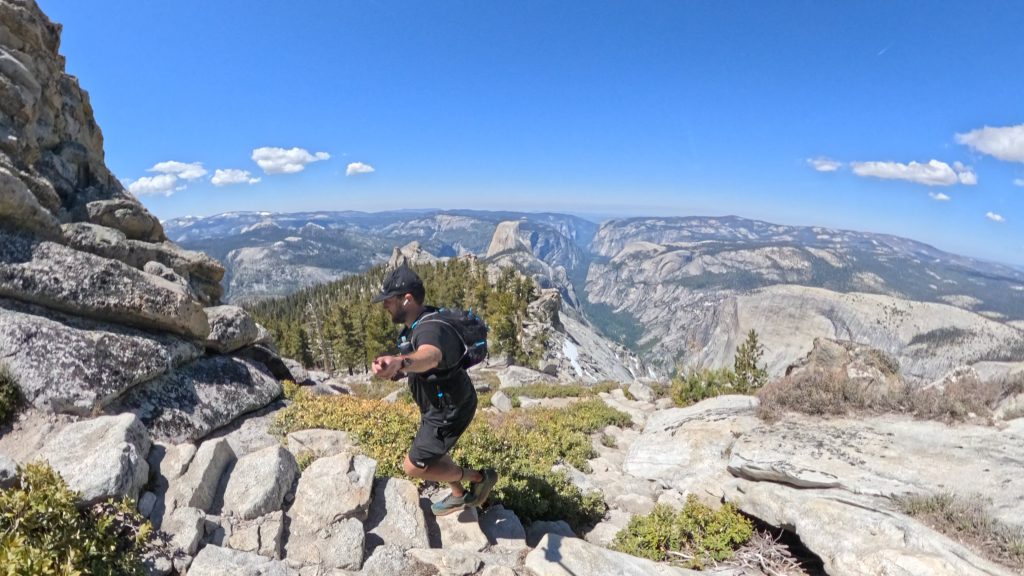 Hiker navigating rocky terrain on the Clouds Rest Trail with Half Dome and Yosemite Valley visible in the background.