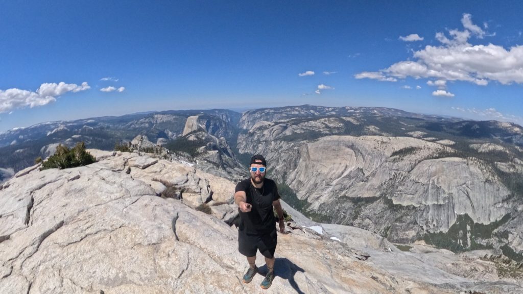 Hiker standing on the summit of Clouds Rest with Yosemite Valley, Half Dome, and granite cliffs in the background.