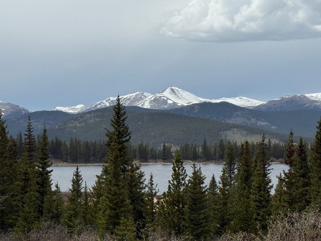 Snow-capped Rocky Mountain peaks rise above evergreen forests and a mountain lake along the Chicago Lakes Trail in Colorado beneath dramatic storm clouds.