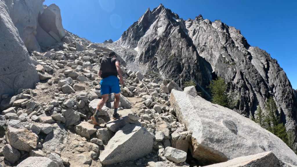 Hiker climbing Asgard Pass trail beneath towering granite cliffs on the Enchantments Traverse in Washington's Alpine Lakes Wilderness.