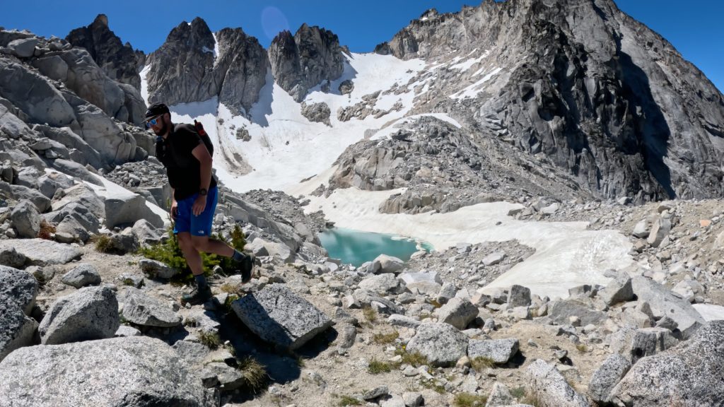 Hiker crossing a rocky slope above a turquoise alpine lake with snowfields and granite peaks during the Enchantments Traverse in Washington.