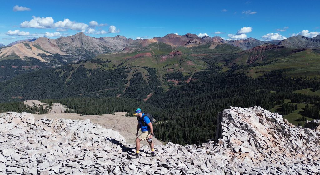 Hiker traversing a rocky summit area on Engineer Mountain with panoramic views of the San Juan Mountains in Colorado