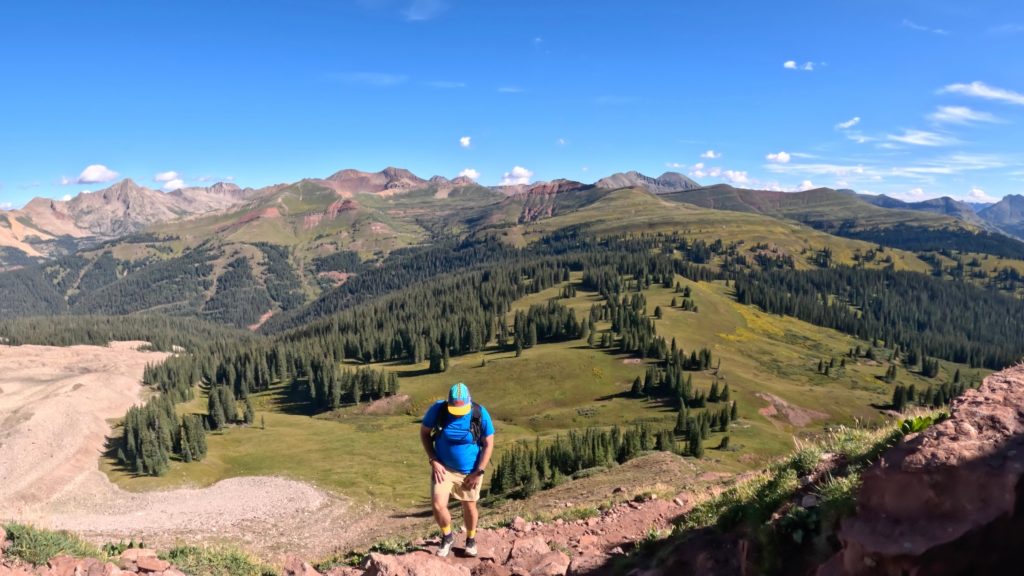 Hiker ascending the Engineer Mountain trail above alpine meadows and evergreen forests in the San Juan Mountains of Colorado.