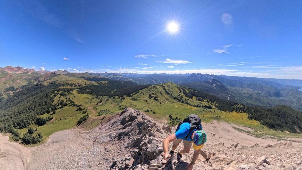 Hiker scrambling up a rocky ridge near the summit of Engineer Mountain with sweeping views of alpine meadows and distant mountain ranges in Colorado.