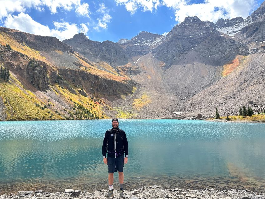 Hiker standing beside the turquoise waters of Lower Blue Lake with rugged San Juan Mountain peaks rising above the alpine basin near Ridgway, Colorado