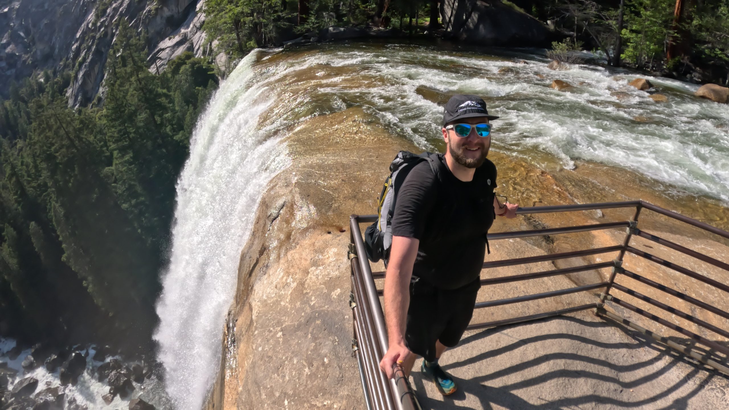Hiker standing at the railing overlooking the top of Vernal Falls in Yosemite National Park on the Mist Trail with rushing water flowing over the granite cliff.