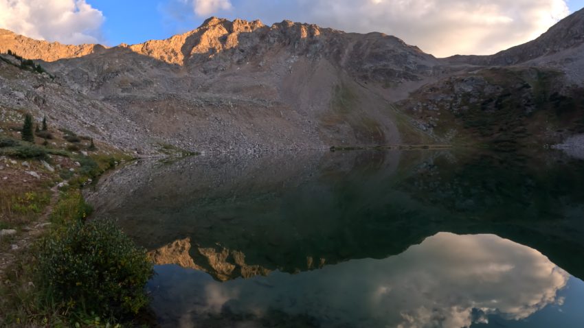 Mountain peaks reflected in the calm waters of Lake Ann during sunset along the Lake Ann Trail in Colorado.