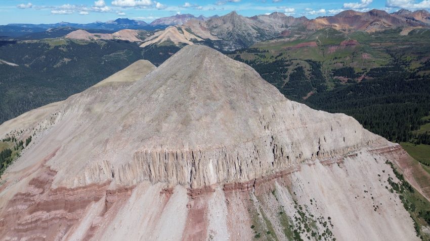 Aerial view of Engineer Mountain's rocky summit ridge with colorful peaks and alpine forests stretching across the San Juan Mountains in Colorado.